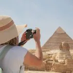 A Tourist woman wearing a sunhat takes photos of the Giza pyramids and the Sphinx in Egypt using a camera.