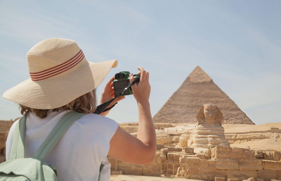 A Tourist woman wearing a sunhat takes photos of the Giza pyramids and the Sphinx in Egypt using a camera.