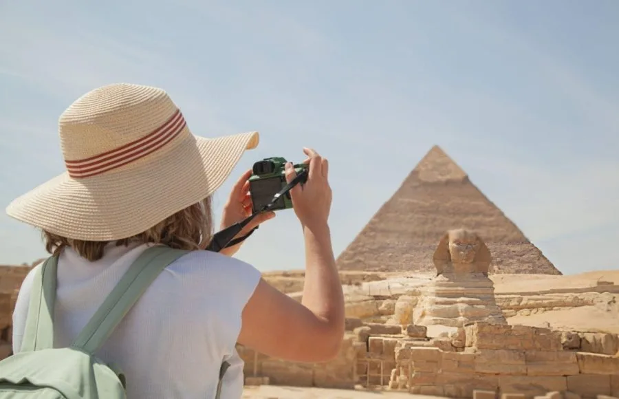 A Tourist woman wearing a sunhat takes photos of the Giza pyramids and the Sphinx in Egypt using a camera.