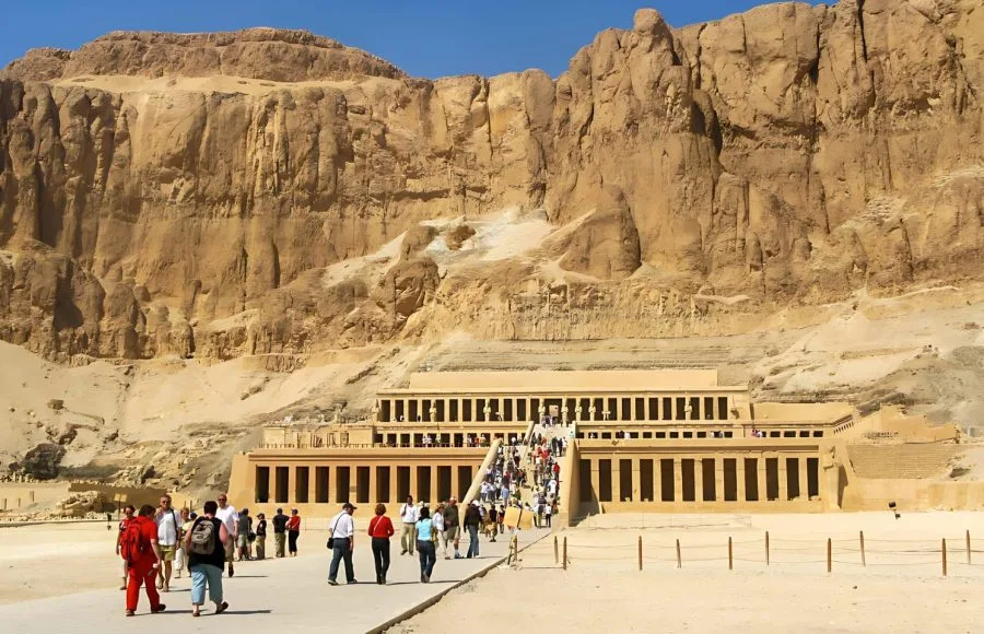 Tourists walk towards the monumental, multi-tiered facade of the hatshepsut temple, built into the towering cliffs on the West Bank of Luxor.