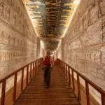 A solo woman tourist walking down a wooden ramp inside a tomb at the Valley of the Kings, with ancient hieroglyphics visible on the walls.