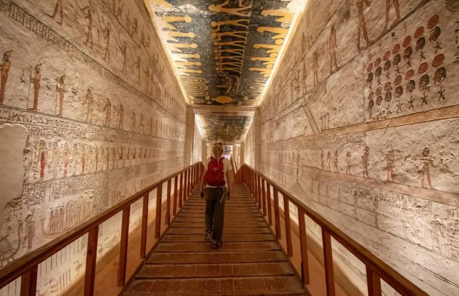 A solo woman tourist walking down a wooden ramp inside a tomb at the Valley of the Kings, with ancient hieroglyphics visible on the walls.