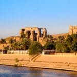 A beautiful view of the ruins of Kom Ombo Temple Egypt on the Nile, showing its columns and exterior walls silhouetted by the sunset over the riverbank and surrounding palm trees.