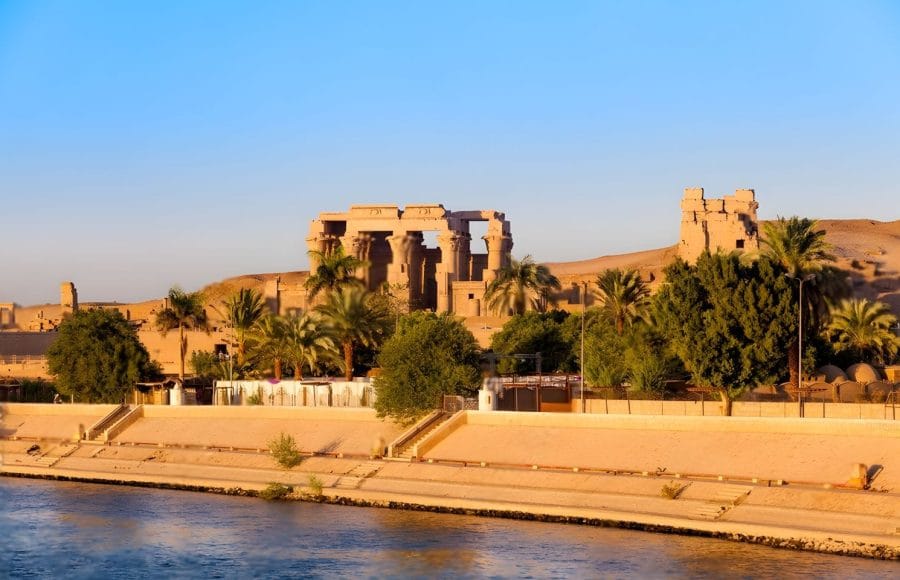 A beautiful view of the ruins of Kom Ombo Temple Egypt on the Nile, showing its columns and exterior walls silhouetted by the sunset over the riverbank and surrounding palm trees.