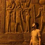 A Solo Traveler stands facing a brightly lit wall of detailed ancient Egyptian carved reliefs at the Temple of Kom Ombo, featuring figures of pharaohs and gods.