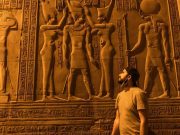 A Solo Traveler stands facing a brightly lit wall of detailed ancient Egyptian carved reliefs at the Temple of Kom Ombo, featuring figures of pharaohs and gods.