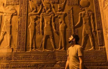 A Solo Traveler stands facing a brightly lit wall of detailed ancient Egyptian carved reliefs at the Temple of Kom Ombo, featuring figures of pharaohs and gods.
