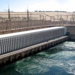 The Aswan High Dam, showing the hydroelectric power station building and the turbulent water flowing from the dam into the Nile River.