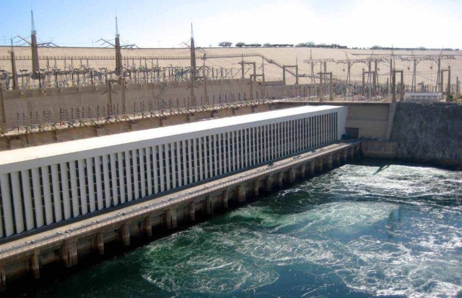 The Aswan High Dam, showing the hydroelectric power station building and the turbulent water flowing from the dam into the Nile River.