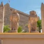 The entrance to The Nubian Museum in Aswan, flanked by ancient Egyptian-style statues and obelisks, set amidst palm trees against a blue sky.