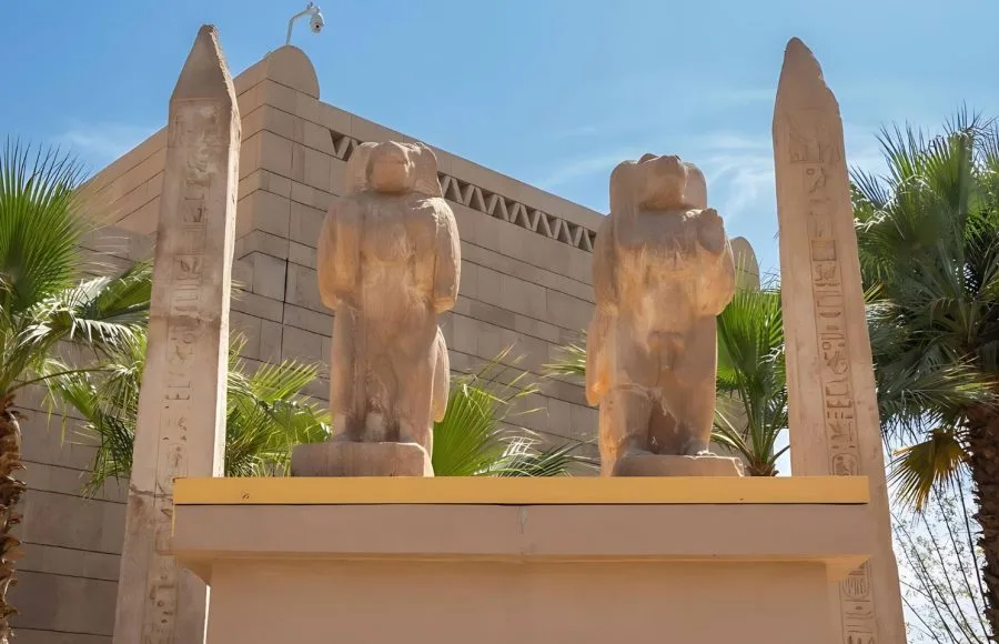 The entrance to The Nubian Museum in Aswan, flanked by ancient Egyptian-style statues and obelisks, set amidst palm trees against a blue sky.