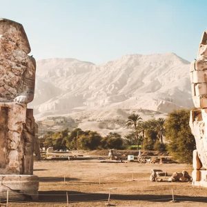 A Photograph Of The Colossi Of Memnon, Two Massive Stone Statues In Egypt Standing Tall Amidst A Barren Desert Landscape.