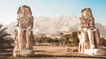 A photograph of the Colossi of Memnon, two massive stone statues in Egypt standing tall amidst a barren desert landscape.
