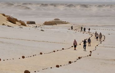 Tourists walk along a desert path marked with stones, leading to domed buildings in a massive, flat, white and sandy landscape on a Fayoum Tour Wadi El Rayan.