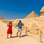 Tourists on camels in the vast desert landscape of the Giza Pyramids, showing the three main large pyramids against a bright blue sky.