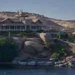 An exterior view of the Aswan Nubian Museum, showing the modern, elegant architecture set into a hillside overlooking the Nile River, with boats docked nearby.