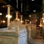 Interior view of the Ben Ezra Synagogue in Coptic Cairo, featuring a marble shrine, decorative columns, and hanging antique brass light fixtures, dimly lit with warm light.