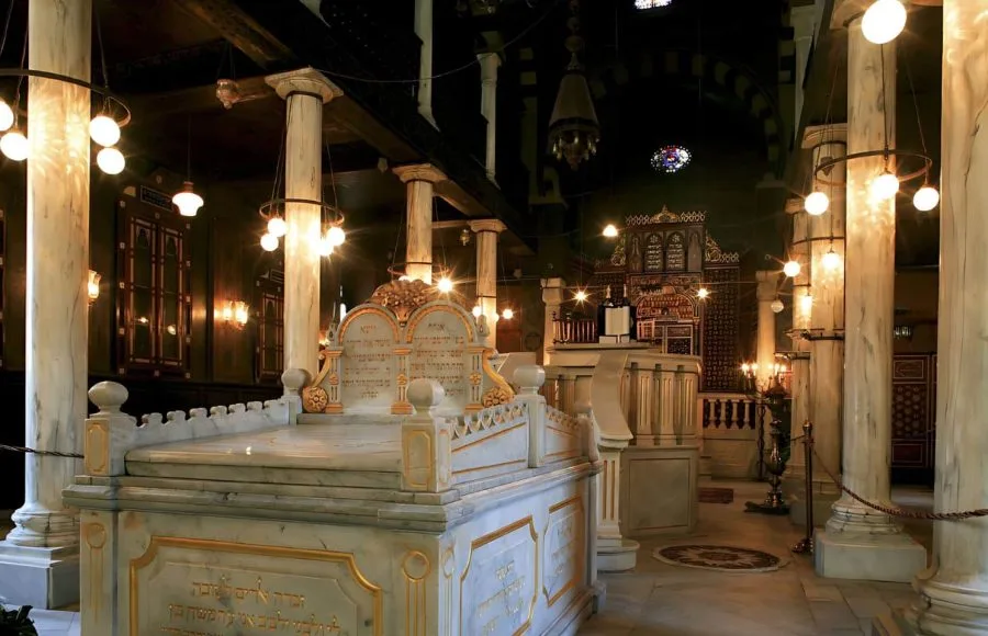 Interior view of the Ben Ezra Synagogue in Coptic Cairo, featuring a marble shrine, decorative columns, and hanging antique brass light fixtures, dimly lit with warm light.