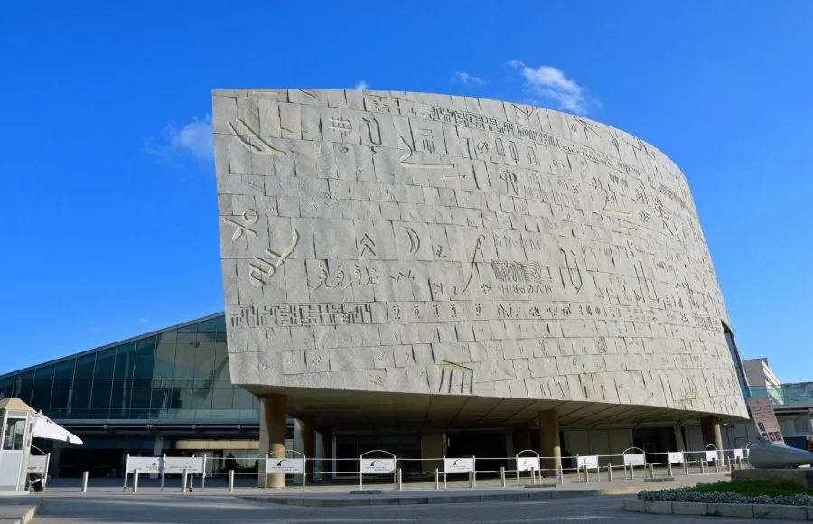The large, curved granite facade of the Bibliotheca Alexandrina in Egypt, covered in carved hieroglyphic-like characters, seen during a daylight Trip.