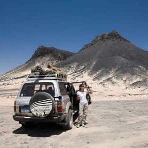A Tourist Standing Next To A Toyota Land Cruiser Suv Taking A Photo In The Black Desert Of Bahariya, With Cone-Shaped Black Volcanic Hills In The Background.