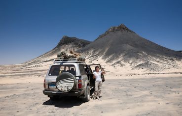 A tourist standing next to a Toyota Land Cruiser SUV taking a photo in the Black Desert of Bahariya, with cone-shaped black volcanic hills in the background.