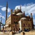 The large, domed structure of the Muhammad Ali Mosque and its tall, slender minarets, situated within the stone walls of the Cairo Citadel against a bright blue, cloudy sky.