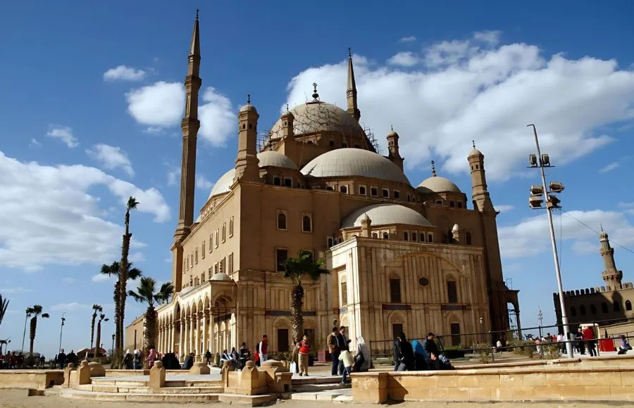 The large, domed structure of the Muhammad Ali Mosque and its tall, slender minarets, situated within the stone walls of the Cairo Citadel against a bright blue, cloudy sky.