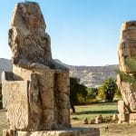 The two colossal statues of Amenhotep III, known as the Colossi of Memnon, sit ruined in a lush field with the mountains of the Theban Necropolis.