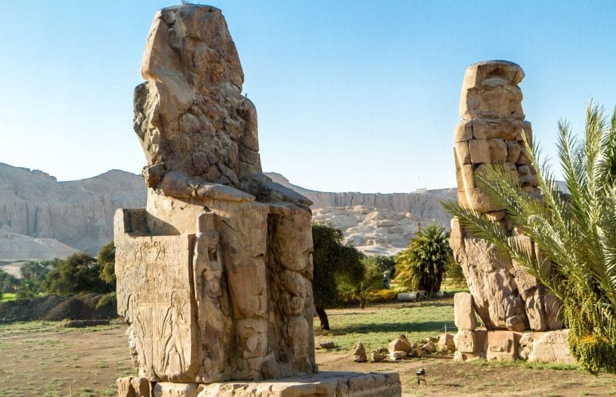 The two colossal statues of Amenhotep III, known as the Colossi of Memnon, sit ruined in a lush field with the mountains of the Theban Necropolis.