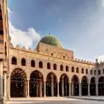 The large, sunlit Courtyard of Mosque of Al Naisr Muhammad inside the Citadel of Cairo, showing its distinctive green dome, arches, and columns.