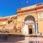 The distinctive salmon-pink facade of the Egyptian Museum building in Tahrir Square, Cairo, featuring a colossal stone sphinx statue in the foreground and flags flying on the roof.