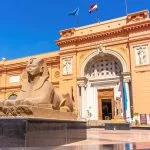 The distinctive salmon-pink facade of the Egyptian Museum building in Tahrir Square, Cairo, featuring a colossal stone sphinx statue in the foreground and flags flying on the roof.