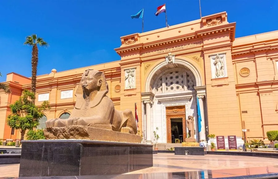The distinctive salmon-pink facade of the Egyptian Museum building in Tahrir Square, Cairo, featuring a colossal stone sphinx statue in the foreground and flags flying on the roof.