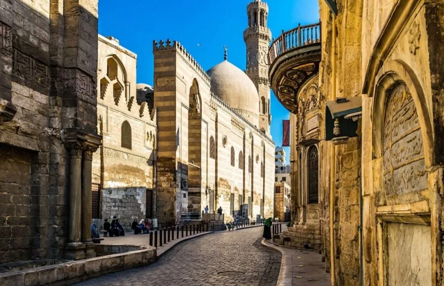The sunlit cobblestone street of El Muizz Street Old Cairo, showing medieval Islamic architecture with domes, arched facades, and a towering minaret.
