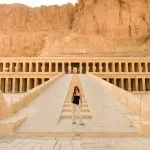 A Female Traveler standing on the central ramp leading up to the multi-tiered Temple of Hatshepsut in Luxor, backed by a sheer cliff face.