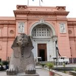 The large, pinkish-orange facade and main entrance of the Egyptian Museum building, with a sphinx statue and water feature in the foreground in Cairo, Egypt.