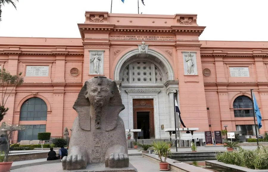 The large, pinkish-orange facade and main entrance of the Egyptian Museum building, with a sphinx statue and water feature in the foreground in Cairo, Egypt.