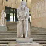 A colossal seated statue of a pharaoh, likely Ramesses II, stands outside the modern, stepped entrance of the Grand Egyptian Museum (GEM) Giza.