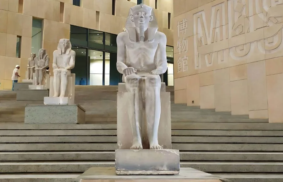 A colossal seated statue of a pharaoh, likely Ramesses II, stands outside the modern, stepped entrance of the Grand Egyptian Museum (GEM) Giza.