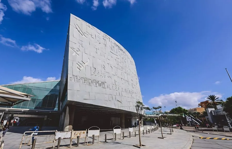 The large, modern, cylindrical exterior facade of the Great Library of Alexandria, Egypt, featuring carved letters and symbols, viewed from a sidewalk under a bright blue sky.