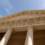 The grand, classical facade of the Greco-Roman Museum in Alexandria, Egypt, featuring tall, smooth columns and a triangular pediment inscribed with Greek letters "ΜΟΥΣΕΙΟΝ" (Mouseion).