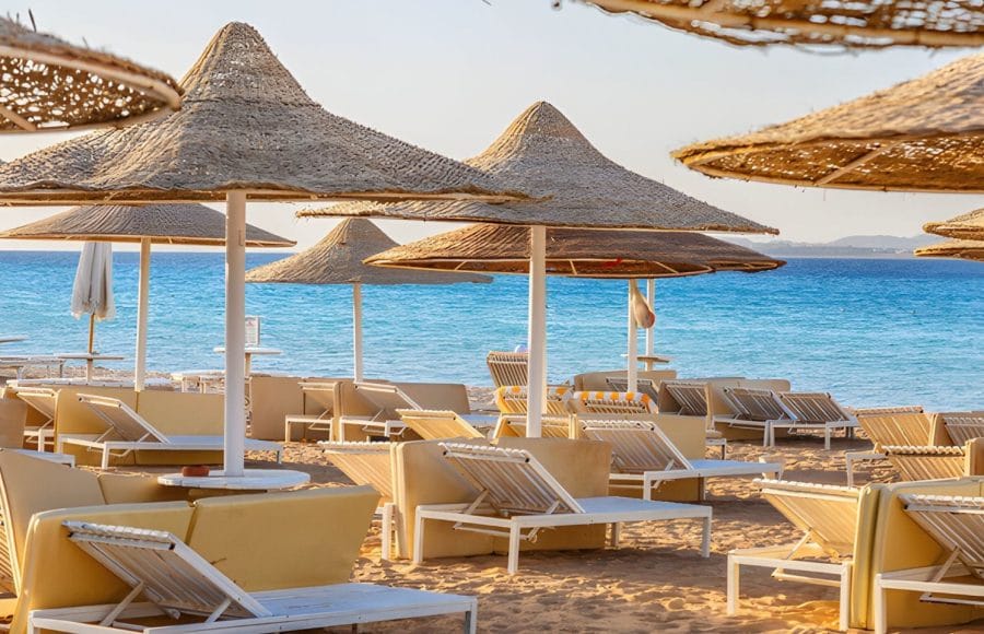 Beach umbrellas and lounge chairs on a golden sandy beach leading to the clear turquoise waters of the Red Sea under a sunny sky.