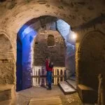 A Tourist stands in the dimly lit, arched underground passage, viewing a chamber Inside Ancient Catacombs of Alexandria, with blue light illuminating the rough-cut stone.