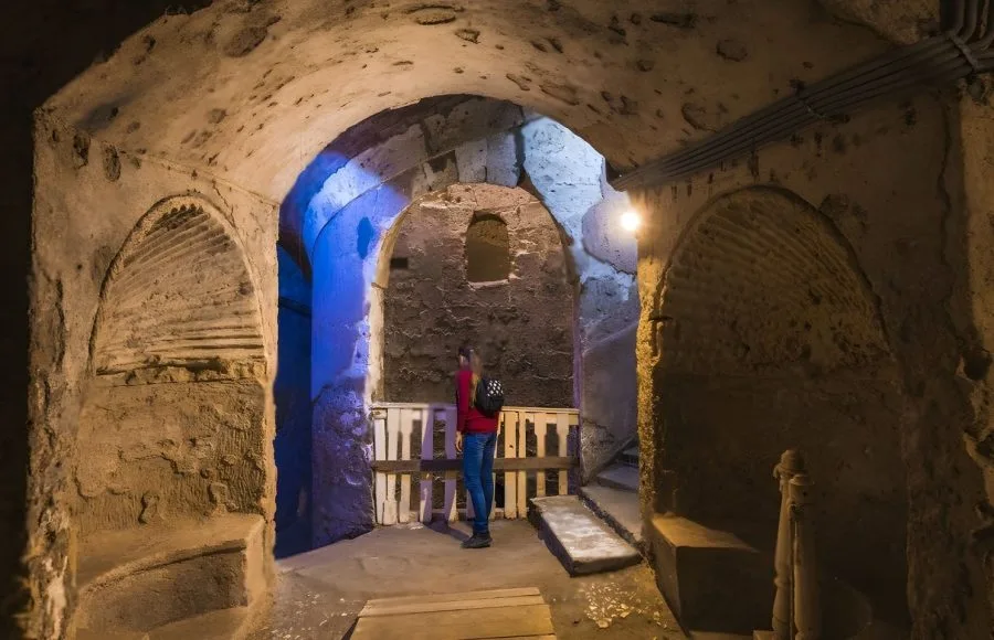 A Tourist stands in the dimly lit, arched underground passage, viewing a chamber Inside Ancient Catacombs of Alexandria, with blue light illuminating the rough-cut stone.