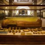 A wide display case Inside Egyptian Museum, Cairo, showing a preserved wooden sarcophagus (coffin) and numerous artifacts, including jars and dishes, set against the dimly lit, vaulted interior of the museum.