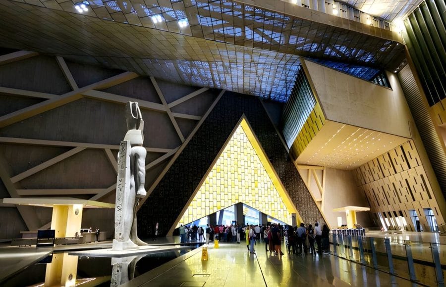 The Grand Egyptian Museum (GEM) welcomes visitors with its spacious, modern interior, featuring a towering statue, and groups of people exploring and enjoying the space.