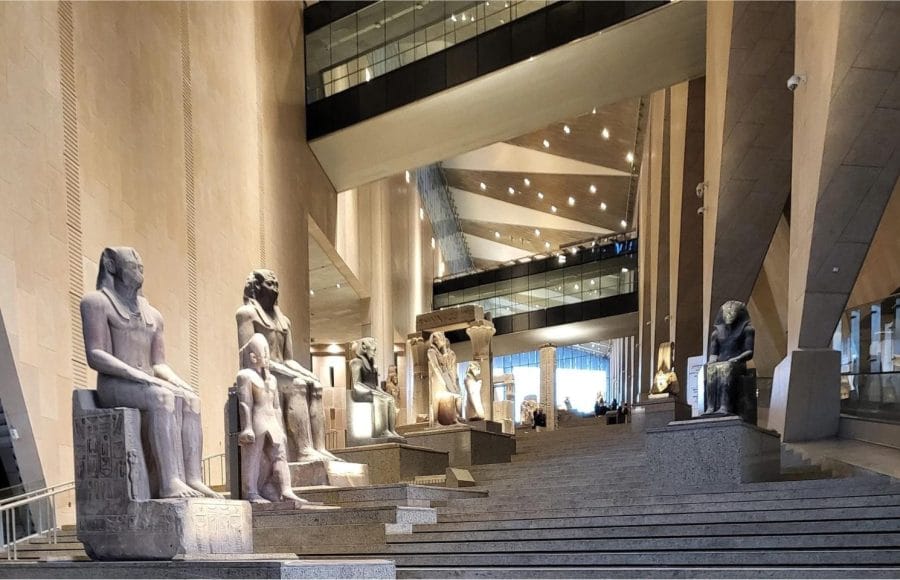 Rows of ancient stone statues of pharaohs and deities displayed on modern steps in the grand, light-filled atrium Inside Grand Egyptian Museum Giza.