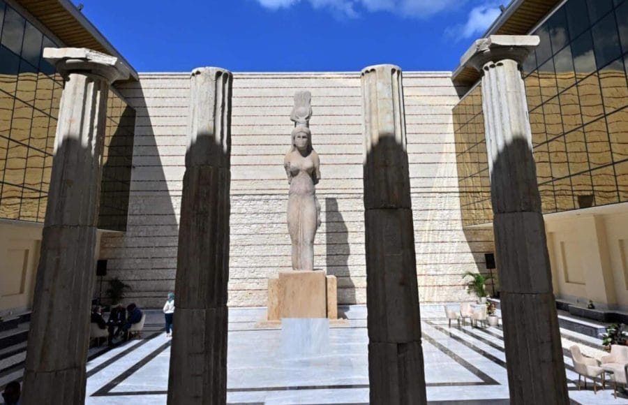 A statue of a draped female figure topped with a modius (headdress) stands in a sunlit courtyard between massive stone columns, showcasing an exhibit Inside Greco-Roman Museum.