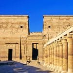 The sunlit main courtyard and facade of the Temple of Philae in Egypt, featuring massive pylons decorated with carved reliefs and a long colonnade with columns on the right.