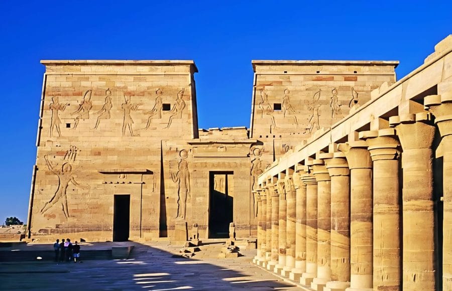 The sunlit main courtyard and facade of the Temple of Philae in Egypt, featuring massive pylons decorated with carved reliefs and a long colonnade with columns on the right.